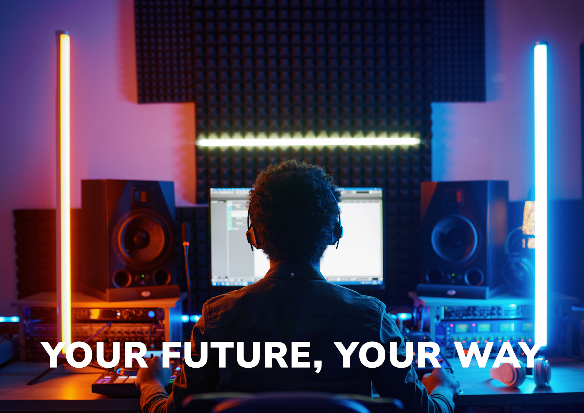 A young person wearing headphones works at a music production workstation with professional studio monitors and colourful LED lighting